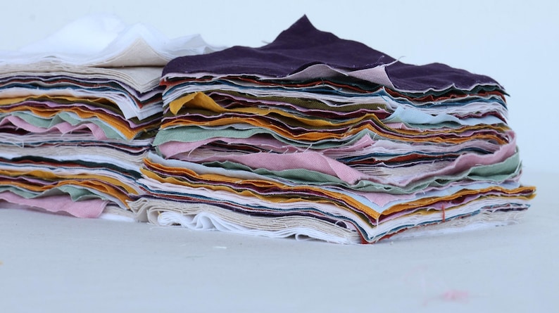 May include: A stack of fabric scraps in various colors, including white, purple, pink, yellow, and green. The fabric is folded neatly and arranged in a pile.