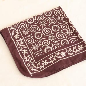 May include: A square, folded bandana in a deep maroon color. The bandana features a repeating white floral and swirl pattern. The border is decorated with a white floral design. The fabric is soft and appears to be cotton.