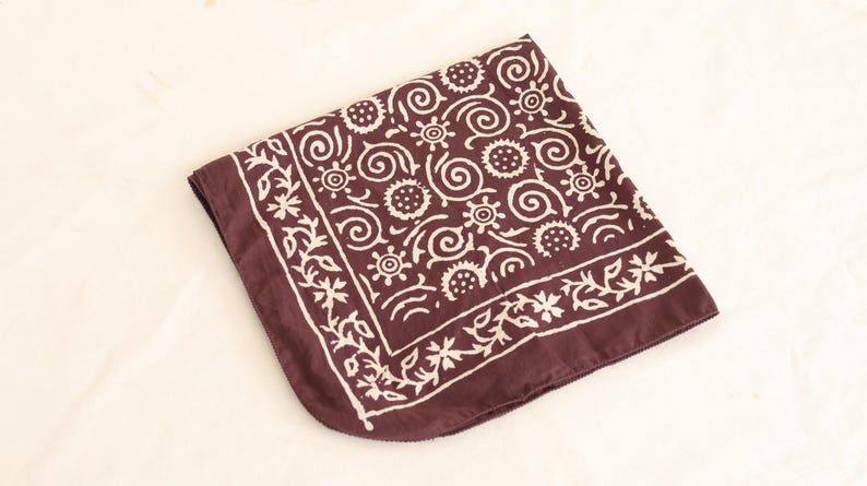 May include: A square, folded bandana in a deep maroon color. The bandana features a repeating white floral and swirl pattern. The border is decorated with a white floral design. The fabric is soft and appears to be cotton.