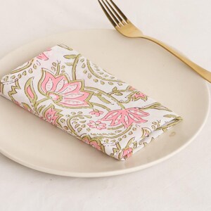 May include: A folded white cloth napkin with a pink and green floral pattern rests on a cream-colored plate. A gold-colored fork is placed to the right of the plate. The napkin features a repeating design of stylized flowers and leaves.