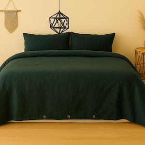 Könnte beinhalten: Ein dunkelgrünes Bettwäsche-Set mit zwei Kissenbezügen und einem Bettbezug. Der Bettbezug hat drei Holzknöpfe am Fußende. Das Bett steht in einem Raum mit neutraler Farbpalette, mit einer dekorativen Wandbehänge und einem Beistelltisch.