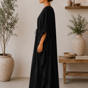 May include: A long, flowing black dress with a V-neck and a drawstring waist. The dress has wide sleeves and a split hem. The person is wearing sunglasses and has their hair up in a bun. The background includes plants and wooden furniture.