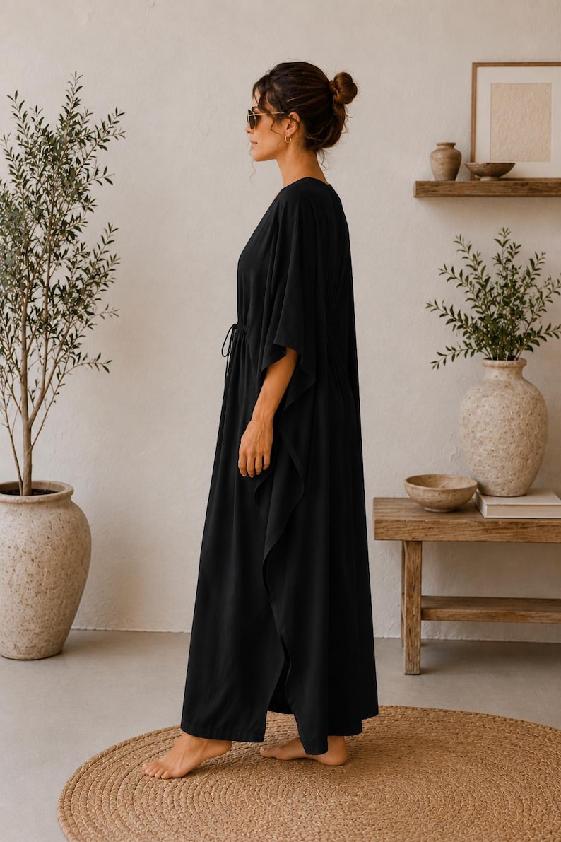 May include: A long, flowing black dress with a V-neck and a drawstring waist. The dress has wide sleeves and a split hem. The person is wearing sunglasses and has their hair up in a bun. The background includes plants and wooden furniture.