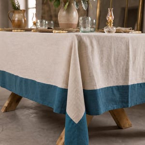Puede incluir: Un mantel rectangular que presenta un panel central de color beige natural con un borde ancho de color azul cerceta sólido a lo largo del borde inferior. La tela tiene una textura tejida visible y cae sobre las esquinas de una mesa de madera.