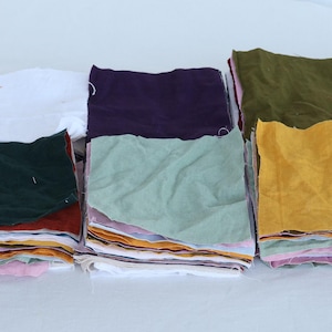 May include: A stack of fabric squares in various colors, including white, green, purple, brown, yellow, and light green. The fabric squares are stacked on top of each other, creating a layered effect.