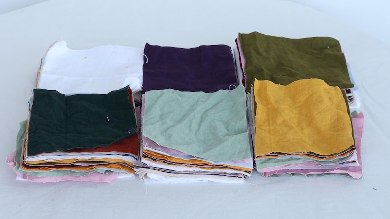 May include: A stack of fabric squares in various colors, including white, green, purple, brown, yellow, and light green. The fabric squares are stacked on top of each other, creating a layered effect.