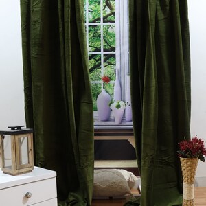 May include: A pair of dark green velvet curtains hanging in front of a window with a nature scene. The curtains are fully drawn and the window is visible through the fabric.
