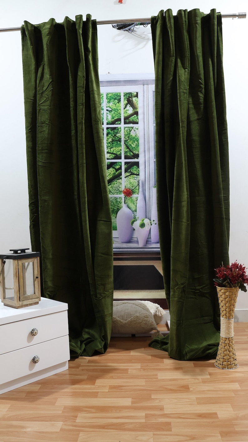 May include: A pair of dark green velvet curtains hanging in front of a window with a nature scene. The curtains are fully drawn and the window is visible through the fabric.