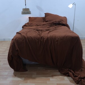 May include: A brown linen duvet cover is neatly arranged on a bed with two pillows. The duvet cover is rumpled and has a relaxed, casual look. The bed is in a room with a white wall and a wooden floor.