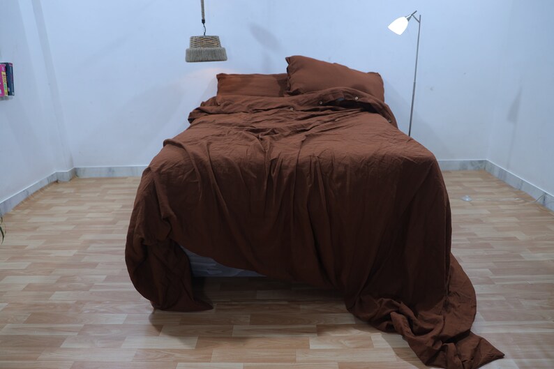 May include: A brown linen duvet cover is neatly arranged on a bed with two pillows. The duvet cover is rumpled and has a relaxed, casual look. The bed is in a room with a white wall and a wooden floor.