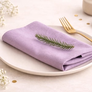 May include: A folded lavender linen napkin with a sprig of greenery rests on a white plate. A gold fork is placed to the side. The scene is set on a light-colored surface, with small white flowers and gold confetti.