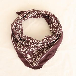 May include: A dark maroon bandana with a white floral and swirl pattern. The square scarf is folded in a triangle shape, with the corners pointing down. The fabric is soft and appears to be cotton.