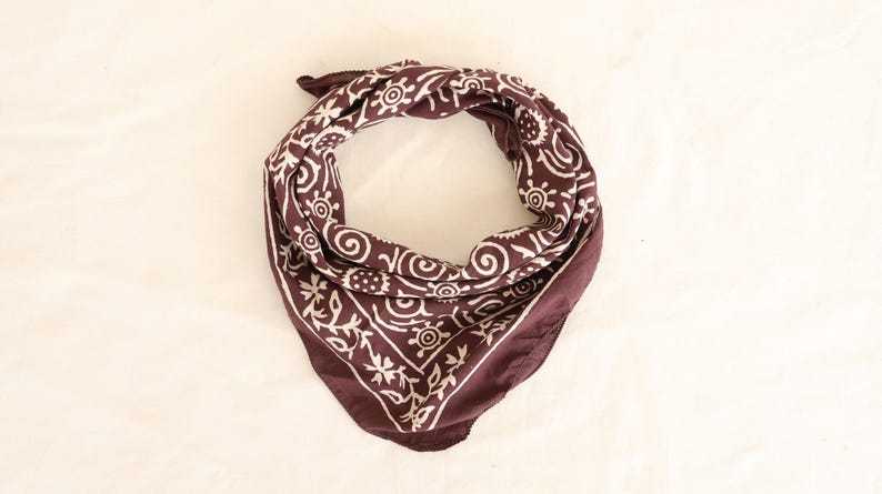 May include: A dark maroon bandana with a white floral and swirl pattern. The square scarf is folded in a triangle shape, with the corners pointing down. The fabric is soft and appears to be cotton.