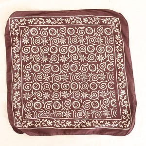 May include: A square, dark brown bandana with a white floral and swirl pattern. The bandana has a decorative border with small flowers. The fabric is soft and appears to be lightweight.