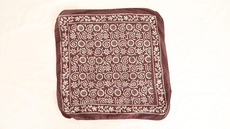 May include: A square, dark brown bandana with a white floral and swirl pattern. The bandana has a decorative border with small flowers. The fabric is soft and appears to be lightweight.