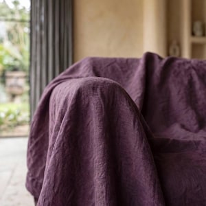 May include: A deep purple fabric throw blanket with a visible woven texture draped over a piece of furniture. The textile features a matte finish and natural folds, showcasing the dense, consistent color and the intricate weave of the material.