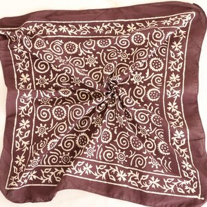 May include: A dark brown square scarf with a white floral and swirl pattern. The scarf is folded and knotted in the center, showcasing the intricate design. The border features a repeating floral motif.