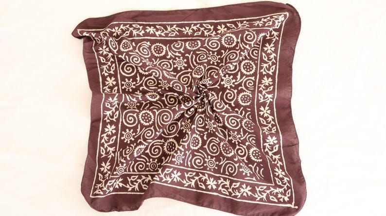 May include: A dark brown square scarf with a white floral and swirl pattern. The scarf is folded and knotted in the center, showcasing the intricate design. The border features a repeating floral motif.