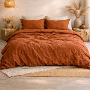 May include: A bedroom scene with a bed dressed in rust-coloured linen bedding, including a duvet cover and two matching pillowcases. The room has a neutral colour palette with woven baskets, dried plants, and a woven pendant light.