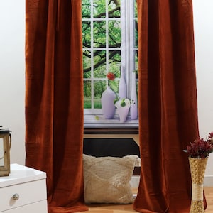 May include: A pair of rust-coloured velvet curtains hang in front of a window with a view of green trees. The curtains are closed and the fabric has a soft, luxurious texture.
