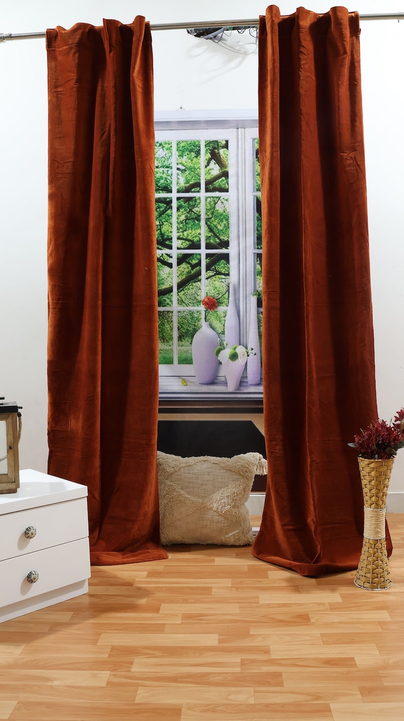 May include: A pair of rust-coloured velvet curtains hang in front of a window with a view of green trees. The curtains are closed and the fabric has a soft, luxurious texture.