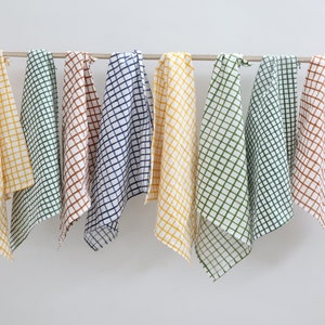 Op de afbeelding: Zeven keukendoeken met een geruit patroon in verschillende kleuren, waaronder geel, groen, bruin en blauw, hangen aan een metalen staaf.