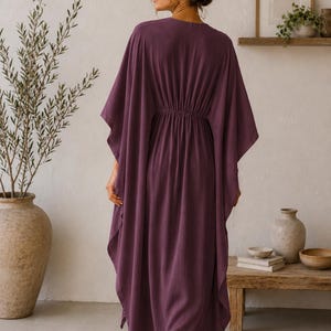 May include: A long, flowing plum-colored caftan dress with a cinched waist. The dress has wide sleeves and a relaxed fit. The model is wearing sunglasses and is standing on a woven rug. The background includes a wooden table and decorative items.
