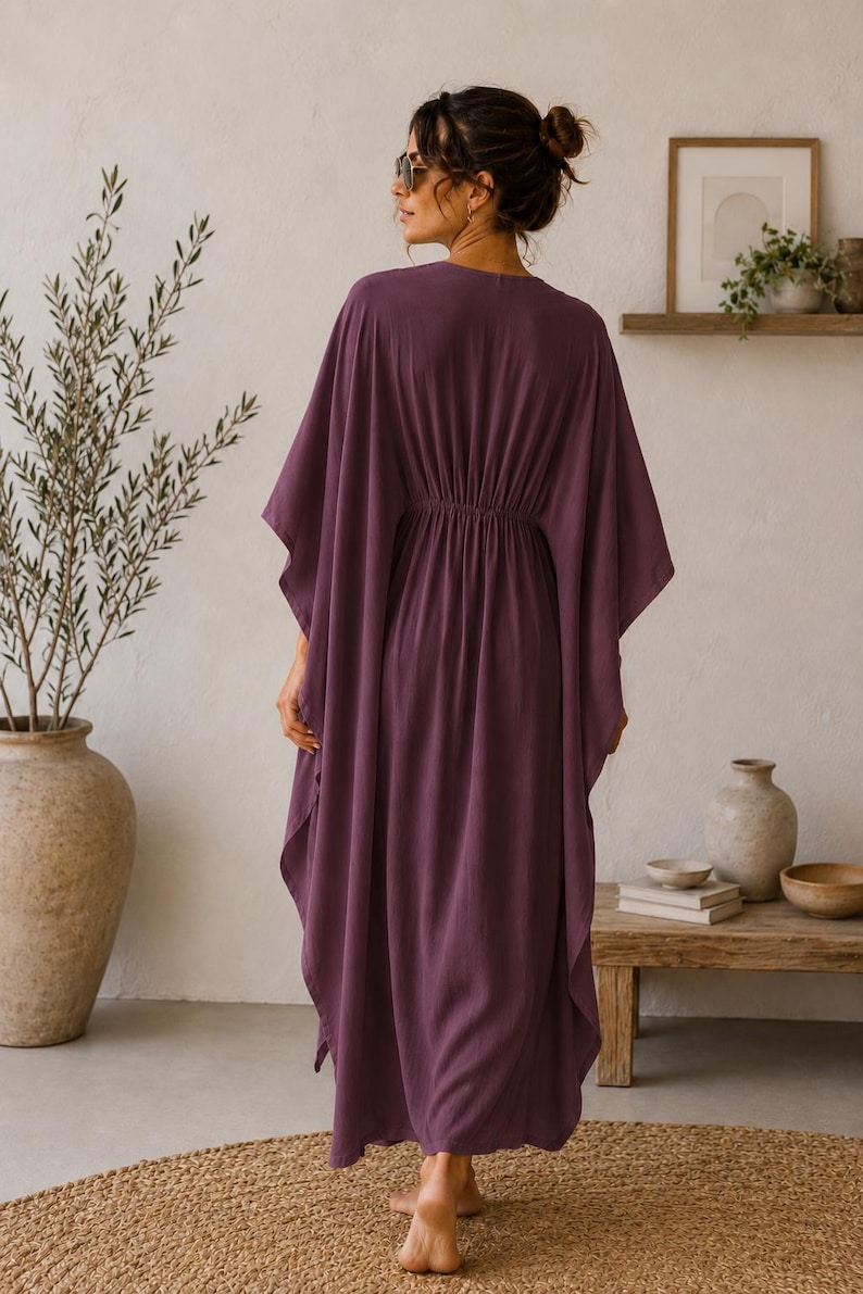 May include: A long, flowing plum-colored caftan dress with a cinched waist. The dress has wide sleeves and a relaxed fit. The model is wearing sunglasses and is standing on a woven rug. The background includes a wooden table and decorative items.