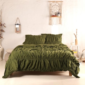 May include: An olive green bedspread and two matching pillowcases are arranged on a wooden bed frame. The bedding has a ruffled design. The room has a white wall and decorative items, including a macrame wall hanging and a wooden lantern.
