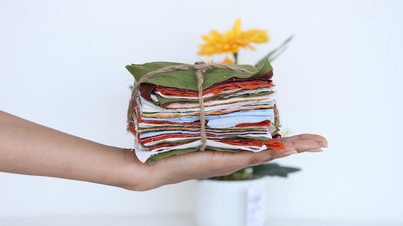 May include: A stack of colorful fabric squares tied together with twine, held in a hand. The fabric squares are in shades of green, brown, orange, red, white, and blue.