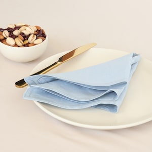 May include: A white ceramic bowl filled with mixed nuts and a light blue linen napkin folded on a white plate with a gold knife.
