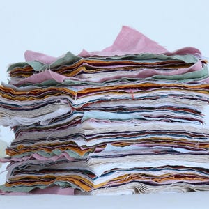 May include: A stack of fabric squares in various colours and patterns, including pink, green, yellow, and white. The fabric squares are stacked on top of each other, creating a colourful and textured pile.