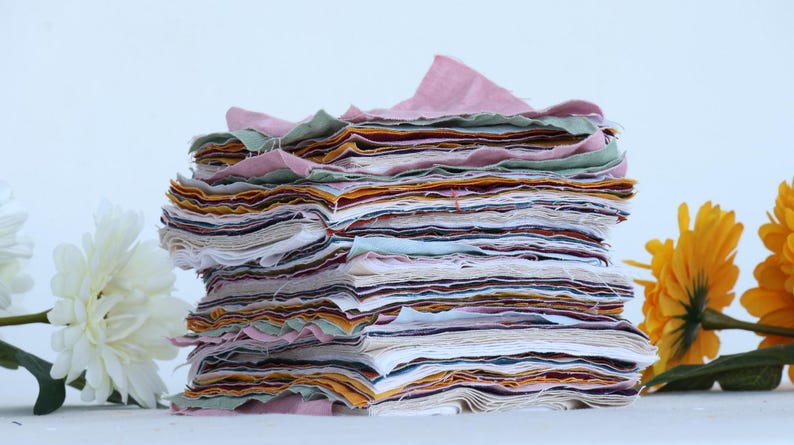 May include: A stack of fabric squares in various colors and patterns, including pink, green, yellow, and white. The fabric squares are stacked on top of each other, creating a colorful and textured pile.