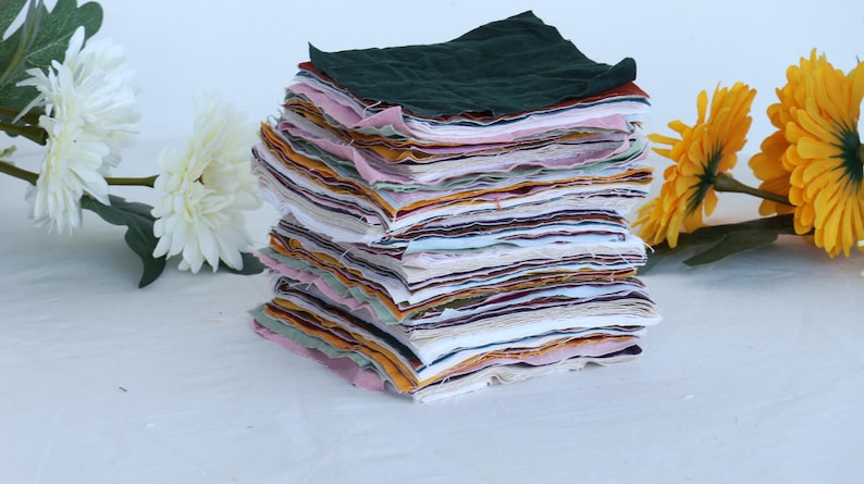 May include: A stack of fabric squares in various colors and patterns, including pink, green, yellow, and white. The fabric squares are arranged in a neat pile, with the top square being a dark green color.