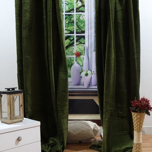 May include: Two dark green velvet curtains hang in front of a window with a view of a green tree and a white windowsill with vases. The curtains are closed and the room has a wooden floor.