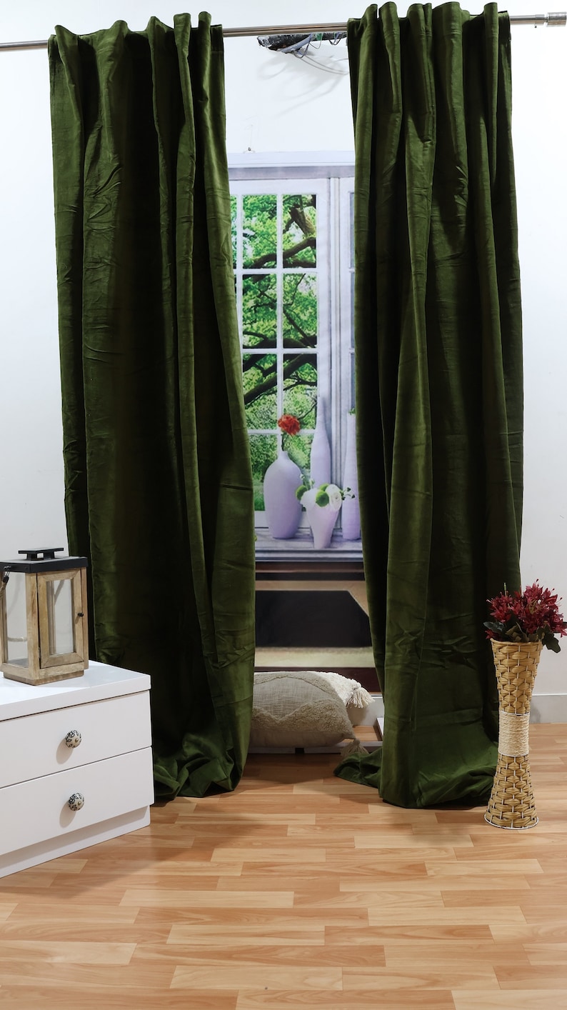 May include: Two dark green velvet curtains hang in front of a window with a view of a green tree and a white windowsill with vases. The curtains are closed and the room has a wooden floor.