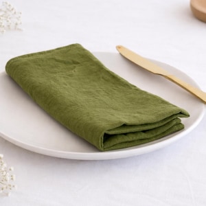 Pode incluir: Uma toalha de linho verde-oliva dobrada repousa sobre um prato branco, com uma faca de manteiga de madeira ao lado. A cena é montada sobre uma toalha de mesa branca, com delicadas flores brancas ao fundo. Tema: decoração de mesa.