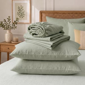 Peut inclure: Un ensemble de literie vert sauge comprenant deux oreillers rectangulaires empilés sur un lit, surmontés d'un drap-housse plié et d'un drap plat. Le tissu présente une texture tissée visible et l'ensemble est présenté dans une chambre claire.