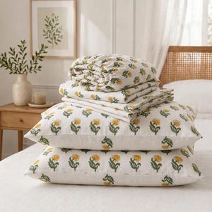 May include: A stack of off-white bedding featuring a repeating pattern of yellow flowers with green stems and leaves. The set includes two rectangular pillows at the base, topped with several folded flat sheets and a fitted sheet.