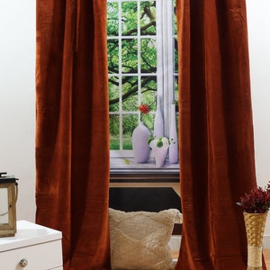 May include: Two rust-coloured velvet curtains hang from a metal rod in front of a window with a view of green trees. The curtains are fully drawn and the window is closed. There is a white cushion on the floor in front of the window.