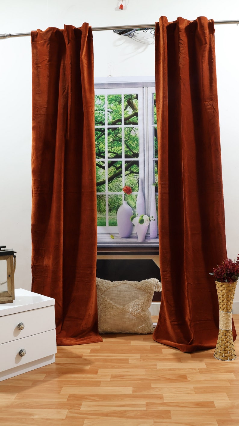 May include: Two rust-coloured velvet curtains hang from a metal rod in front of a window with a view of green trees. The curtains are fully drawn and the window is closed. There is a white cushion on the floor in front of the window.