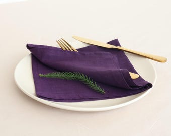 May include: A purple linen napkin folded on a white plate with gold cutlery and a sprig of greenery.