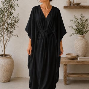 Può includere: Un abito caftano nero e fluido con scollo a V e vita con coulisse. L'abito ha maniche ampie e arriva alle caviglie. La persona indossa occhiali da sole. Lo sfondo comprende un tappeto intrecciato, piante e mensole in legno.