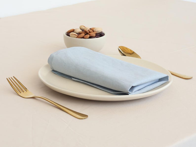 May include: A light blue linen napkin folded on a white plate with a bowl of nuts and two gold utensils.