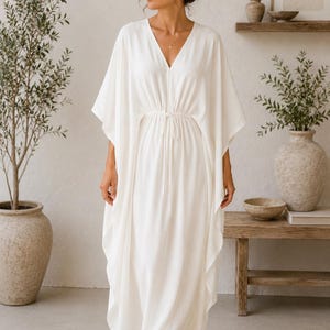 Può includere: Un lungo abito bianco con scollo a V e vita con coulisse. L'abito ha maniche ampie e una vestibilità ampia. La persona indossa occhiali da sole. Lo sfondo mostra un interno dai toni neutri con piante e mobili in legno.