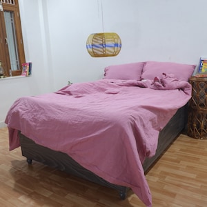 May include: A pink bed with a wooden frame and a woven lampshade hanging above it. The bed is made with a pink duvet and two white pillows. A small wooden side table with a book on top is next to the bed.