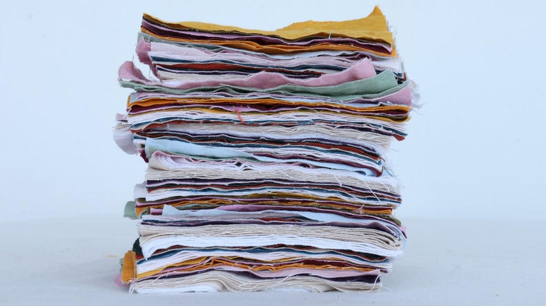 May include: A stack of fabric scraps in various colors, including white, pink, yellow, brown, and blue. The fabric is layered and folded, creating a textured and colorful display.