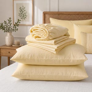 May include: A set of pale yellow bedding including two rectangular pillows stacked on top of each other, topped with a folded flat sheet and a fitted sheet. The fabric has a visible woven texture, and the entire set is presented in a soft, light yellow color palette.