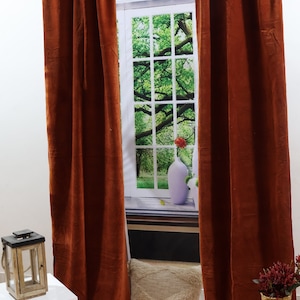 May include: Two rust-coloured velvet curtains hang from a silver curtain rod in front of a window with a green tree and a white vase with a red flower. A white bedside table with two drawers is on the left side of the image. A wooden lantern is on top of the bedside table. A white throw pillow is on the floor in front of the window. A wicker vase with red flowers is on the right side of the image.