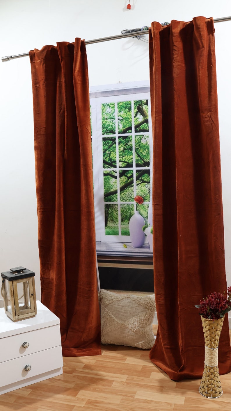 May include: Two rust-coloured velvet curtains hang from a silver curtain rod in front of a window with a green tree and a white vase with a red flower. A white bedside table with two drawers is on the left side of the image. A wooden lantern is on top of the bedside table. A white throw pillow is on the floor in front of the window. A wicker vase with red flowers is on the right side of the image.